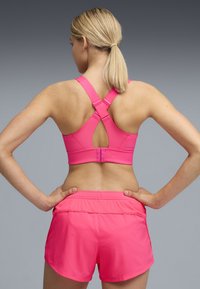 Γυναίκα με ξανθά μαλλιά σε αλογοουρά, που φοράει ροζ αθλητικό σουτιέν με σταυρωτούς ιμάντες και ροζ αθλητικό σορτς που ταιριάζουν, με τα χέρια στη μέση, γυρισμένη προς τα πίσω.