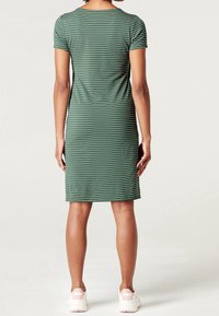 Zelené pruhované šaty s krátkým rukávem viděné zezadu, s těsným střihem a jemnou texturou. Nosené s světlými teniskami.