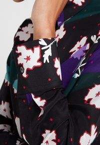 Zwarte bloes met rode en witte bloemen, voorzien van een gladde textuur, een manchetknoop en kleurrijke geometrische accenten in groen en paars.