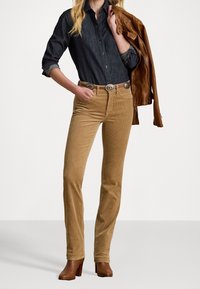 Pantalons en velours côtelé marron associés à une chemise en denim foncé et une veste en cuir marron. Comprend une ceinture à boucle en argent et des bottes à talons marron.