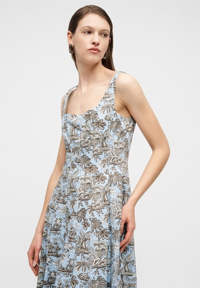 Γυναίκα με καστανά μαλλιά που φορά χρυσούς κρίκους στα αυτιά και ένα αμάνικο, ανοιχτό μπλε φόρεμα με καφέ λουλουδάτο και φυλλώδες μοτίβο.