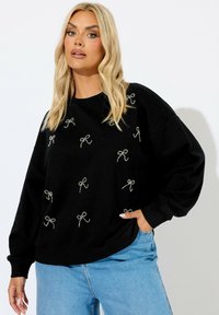 Sudadera negra con bordado de lazo plateado, corte relajado, puños anchos y escote redondo, combinada con jeans de color azul claro.