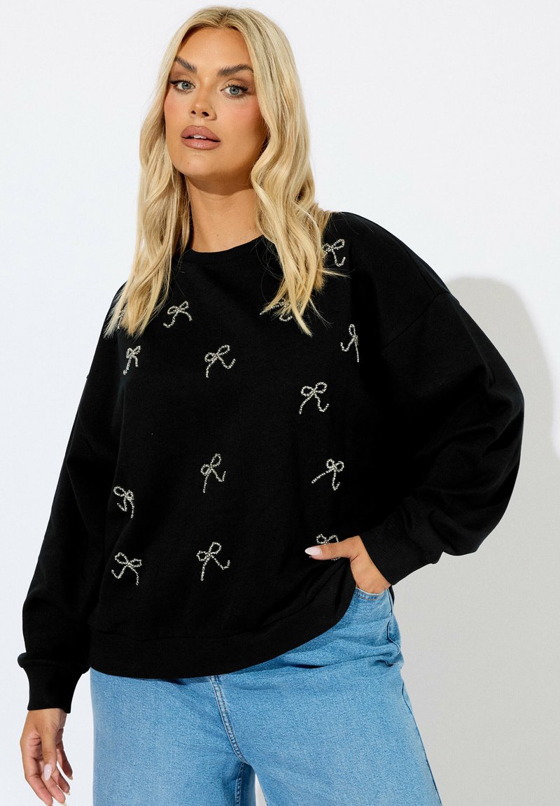 Sudadera negra con bordado de lazo plateado, corte relajado, puños anchos y escote redondo, combinada con jeans de color azul claro.