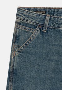 Uvoľnené džíny z denimu v stredne modrej farbe. Majú oranžové prešívanie, tradičný dizajn s piatimi vreckami a predné zapínanie na gombík.