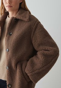 Veste en polaire marron avec une surface texturée, un grand col et des boutons noirs. Le design comprend des poches latérales et une coupe décontractée.