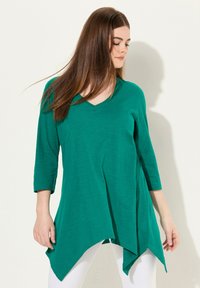 Grünes V-Ausschnitt-Tunika mit dreiviertel Ärmeln, asymmetrischem Saum, aus weichem Stoff mit glatter Textur. Getragen über weißen Leggings.