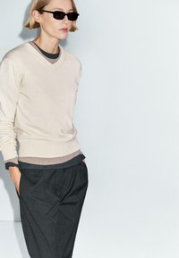 Maglione beige a scollo a V sovrapposto a una maglietta grigio scuro, abbinato a pantaloni scuri. Gli accessori includono occhiali da sole neri. Texture del tessuto liscia.