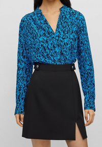 Kvinna klädd i en blåmönstrad långärmad blus instoppad i en svart högmidjad kjol med slits framtill och dekorativa knappar på midjebandet.