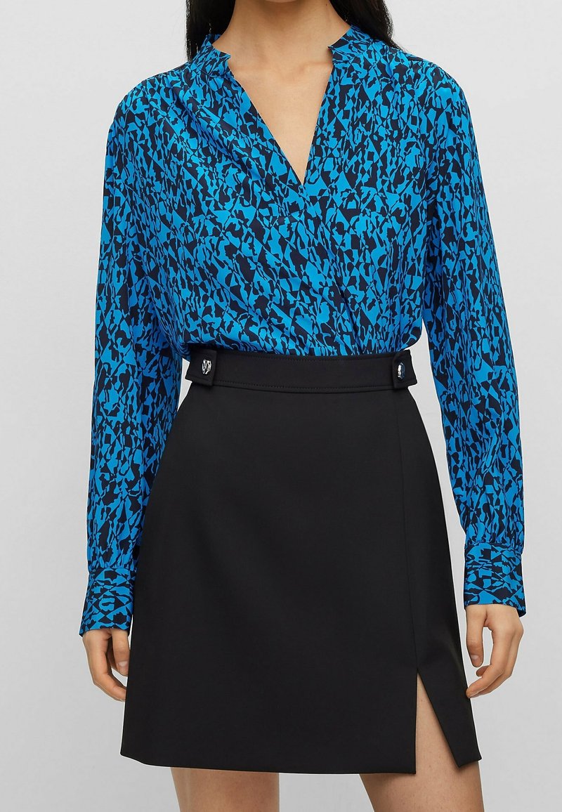 Kvinna klädd i en blåmönstrad långärmad blus instoppad i en svart högmidjad kjol med slits framtill och dekorativa knappar på midjebandet.