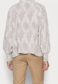 Blouse à manches ballons longues, en tissu crème avec un motif abstrait noir, col boutonné et poignets froncés, associée à un pantalon beige.