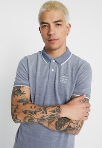 Marineblå polo shirt med hvide detaljer, struktureret stof, korte ærmer og broderet logo på brystet. Tatoveringer synlige på armene.