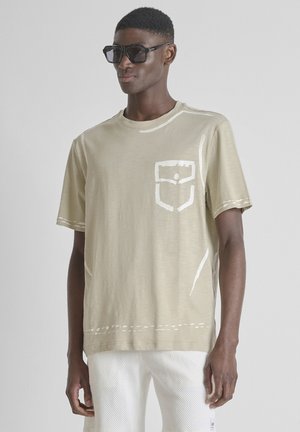 Uomo che indossa una maglietta beige oversize con motivo bianco stampato sulla tasca e pantaloni bianchi texturizzati, accessoriato con grandi occhiali da sole neri.