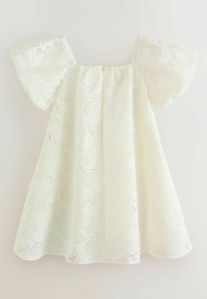 Robe blanche en dentelle à épaules dénudées avec des manches courtes bouffantes et des motifs floraux, présentée sur un fond uni.