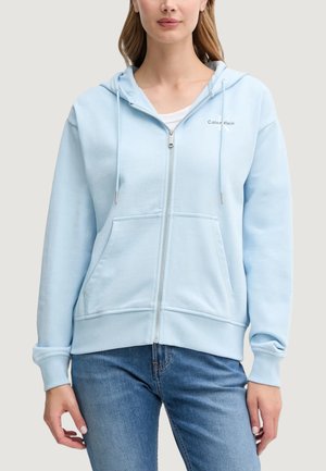 Sudadera con cremallera de color azul claro, con capucha, bolsillos frontales y puños de canalé, que presenta un pequeño logo blanco en el lado izquierdo del pecho. Textura de tela suave.