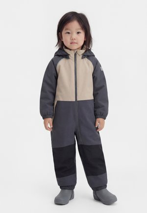 Μικρό παιδί που στέκεται, φοράει δίχρωμη ολόσωμη φόρμα με κουκούλα για εξωτερικούς χώρους με ελαστικές μανσέτες και γκρι μπότες, μπροστά σε απλό φόντο.