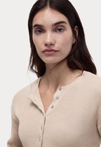 Jasnobeżowy pr�ążkowany top henley z okrągłym dekoltem i trzyguzikowym zapięciem, charakteryzujący się miękką teksturą i dopasowanym krojem.