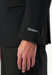 Ręka osoby w czarnym żakiecie z czterema czarnymi guzikami i metką "KARL LAGERFELD" na mankiecie.