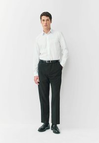 Weißes Hemd mit Knöpfen, schwarze Anzughose, schwarzer Gürtel und schwarze Schuhe, vor einem schlichten weißen Hintergrund stehend.