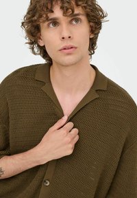 Knit olive green button-up shirt with a textured pattern, collar, and short sleeves; model adjusts the collar with a hand.