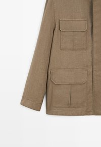 Braune Langarmjacke mit zwei großen Fronttaschen mit Klappe und sichtbaren Nähten vor einfarbig hellem Hintergrund.