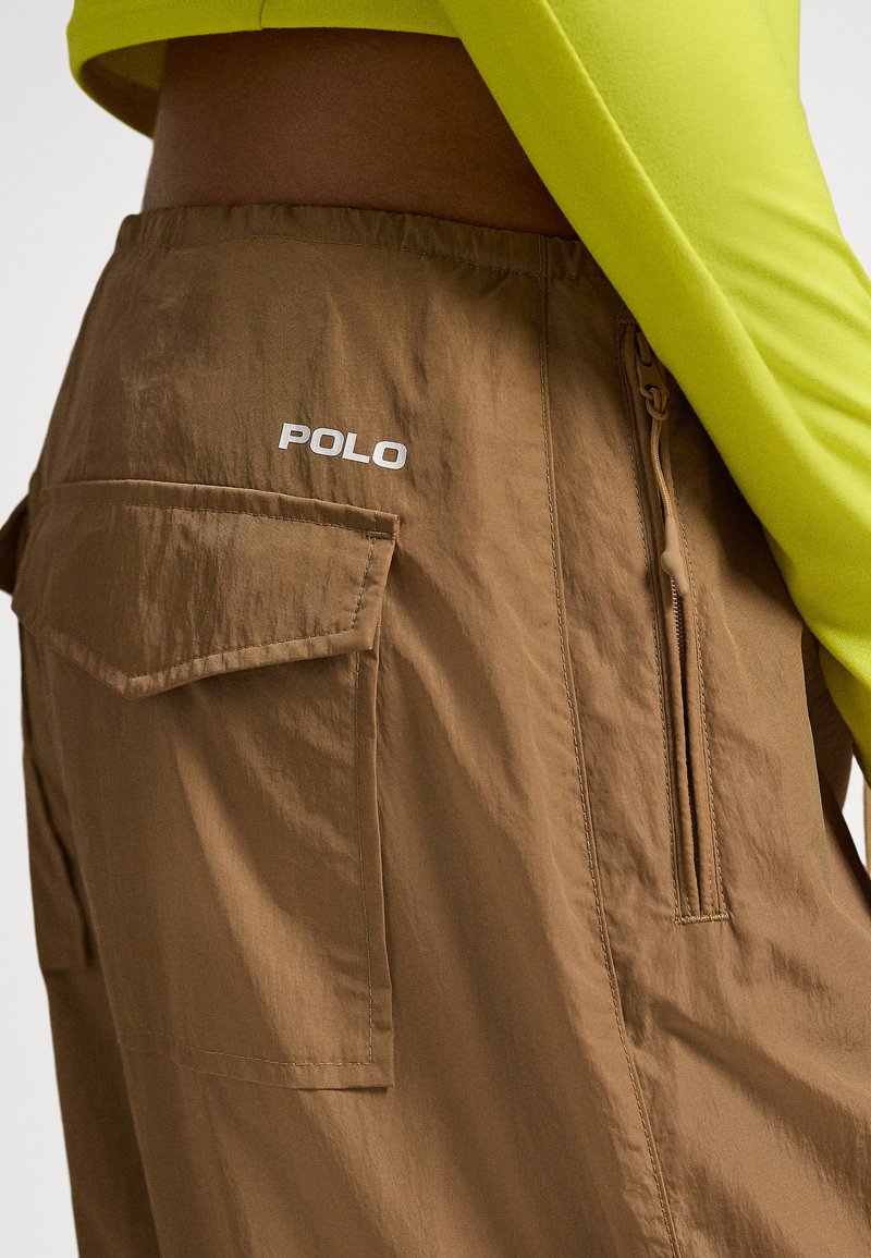 Braune Cargo-Hose mit Reißverschlusstasche und Klappentasche, mit weißem "POLO"-Logo auf der Rückseite, getragen mit einem limettengrünen, kurzen Oberteil.