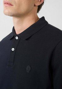 Polo nero realizzato in tessuto testurizzato, con colletto a tre bottoni, bottoni bianchi e una patch nera con logo sul petto.