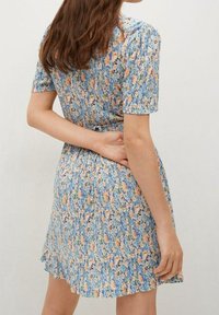 Blumen Kleid in Hellblau mit kurzen Ärmeln. Hat eine geraffte Taille, einen Rückenknoten und ein Muster aus orangefarbenen und grünen Blumen mit strukturierter Oberfläche.
