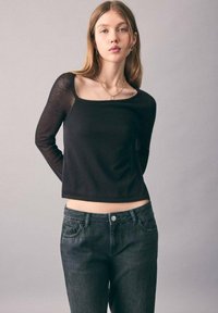 Junge Frau mit langen hellbraunen Haaren, die ein schwarzes Langarmoberteil und dunkelgraue Jeans trägt und vor einem hellgrauen Hintergrund steht.