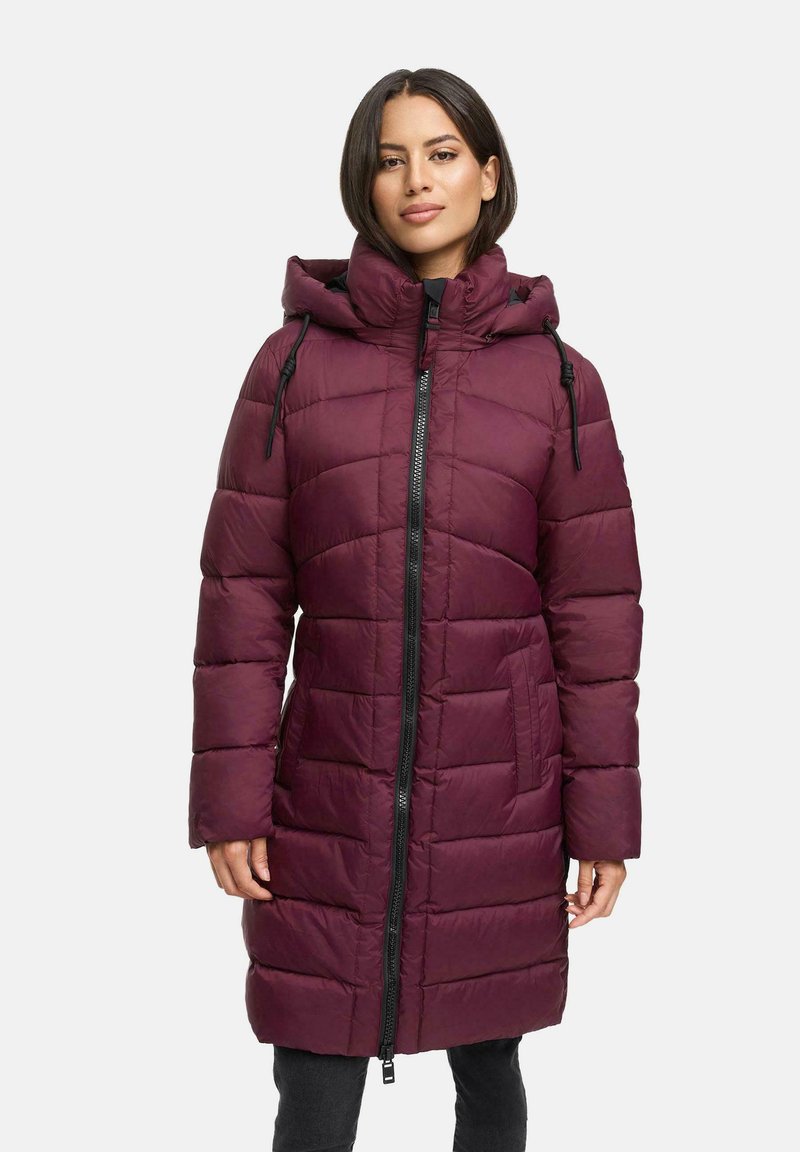 Mujer vestida con un abrigo de invierno acolchado, de longitud a la rodilla, color marrón burdeos, con capucha y cremallera frontal negra, de pie contra un fondo claro y liso.