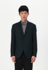 Dunkelgrüner Blazer mit glatter Textur, zwei Knöpfen und zwei Fronttaschen. Über einem grauen Hemd getragen, mit einem klassischen Kragen und Knopfdetails.