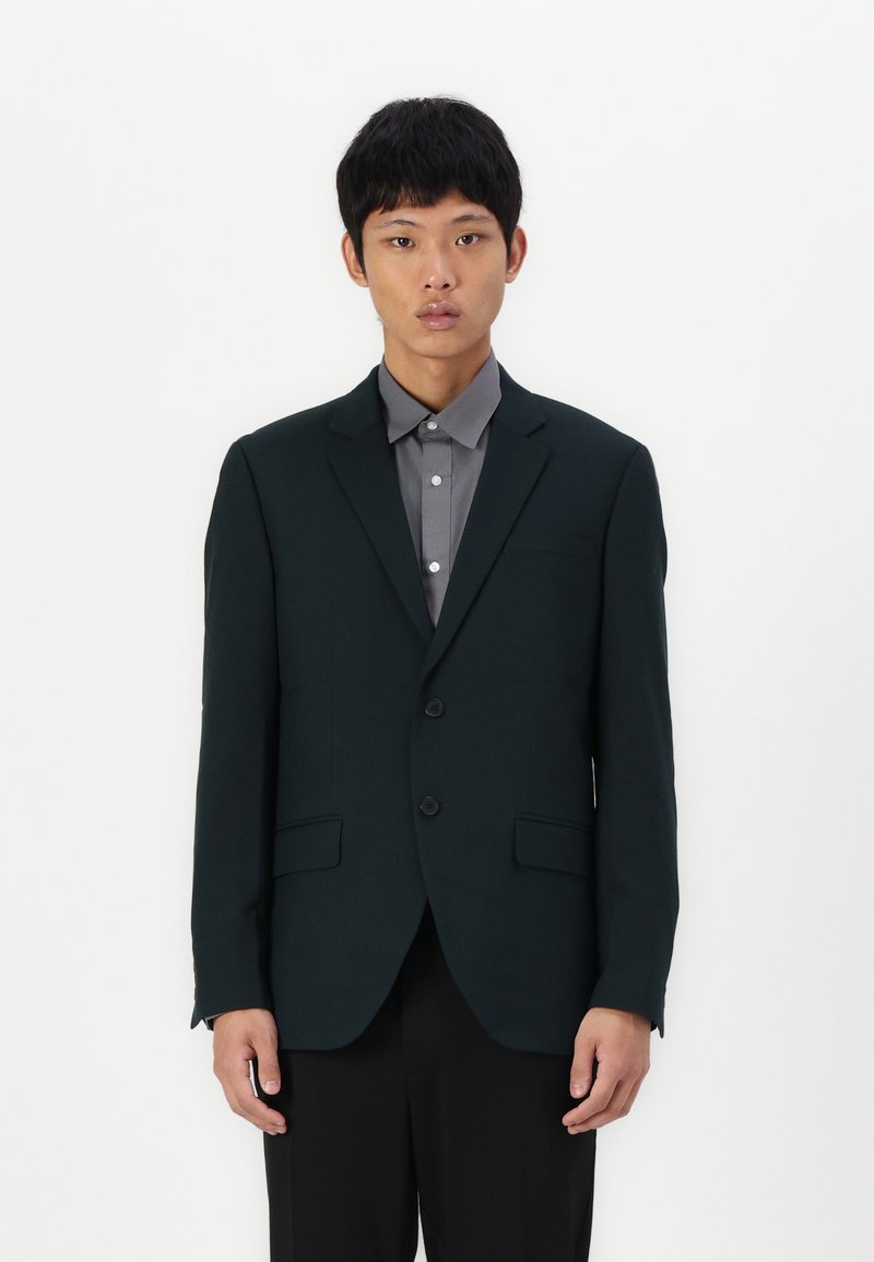 Dunkelgrüner Blazer mit glatter Textur, zwei Knöpfen und zwei Fronttaschen. Über einem grauen Hemd getragen, mit einem klassischen Kragen und Knopfdetails.