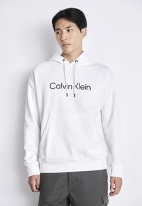Witte hoodie met een voorzak, een trekkoordcapuchon en een zwart "Calvin Klein" logo op de borst. Glad materiaal, casual ontwerp.