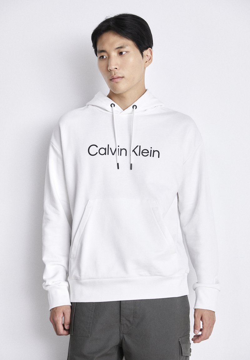 Witte hoodie met een voorzak, een trekkoordcapuchon en een zwart "Calvin Klein" logo op de borst. Glad materiaal, casual ontwerp.