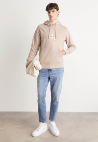 Felpa in cotone beige con logo rosso "Calvin Klein Jeans", tasca frontale, abbinata a jeans azzurri e sneakers bianche, con borsa beige.