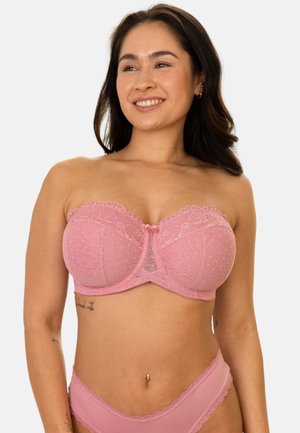 Woman smiling, wearing a pink strapless lace bra and matching underwear, standing against a plain white background.
