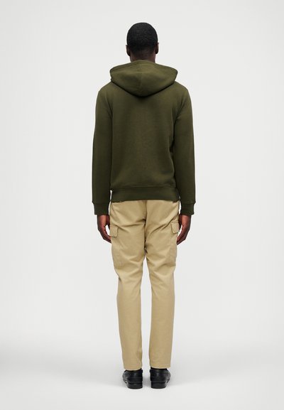 Sweatshirt verde-azeitona com capuz, combinado com calças cargo bejs claras com bolsos, contra um fundo branco liso.