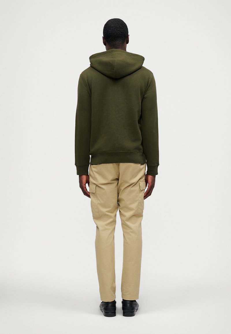 Sudadera verde oliva con capucha, combinada con pantalones cargo beige claros que tienen bolsillos, sobre un fondo blanco liso.