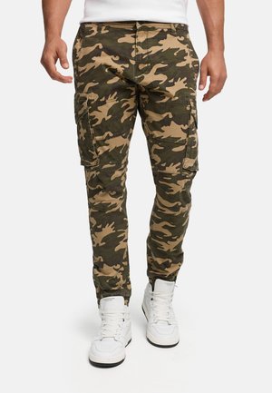 Man wearing green and brown camouflage cargo pants and white high-top sneakers standing against a white background.