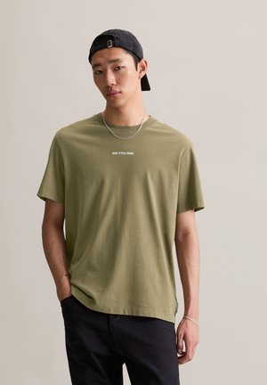 Oliven grønn bomulls T-skjorte med rund hals, korte ermer og trykt logo "MAR C O'POLO DENIM" over brystet. Modellen har på seg en svart caps.