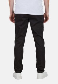 Schwarze Hosen mit geradem Schnitt, aus einem strukturierten Stoff, getragen mit einem weißen Hemd und weißen Sneakers, von hinten betrachtet.