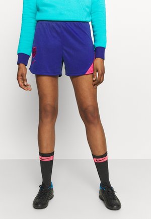 Persona vestida con pantalones cortos deportivos azules y rosas, sudadera turquesa, calcetines Nike negros y rosas, y zapatos de fútbol negros de pie sobre un fondo blanco.