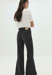 Femme aux longs cheveux foncés se tient de profil, portant un t-shirt blanc rentré dans un jean noir à taille haute et large.