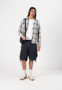 Rutete flannel skjorte i grøn og blå, hvid t-shirt, mørke cargo shorts med sidelommer, hvide sneakers med gummisål, sort crossover taske.