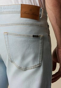 Světle modré denimové džíny s texturovanou tkaninou, designem se pěti kapsami a hnědou koženou nášivkou na zadní straně s embosovaným textem.