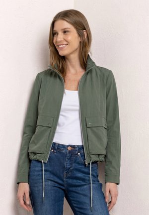 Kvinde smiler, iført en grøn lynlåstrøje med snore og klaplommer over en hvid top og blå jeans, stående mod en tekstureret væg.
