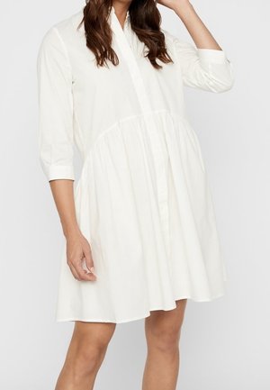 Kobieta w luźnej, białej sukience do kolan z rękawami do połowy przedramienia i marszczoną spódnicą, stoi z uniesionym jednym ramieniem.