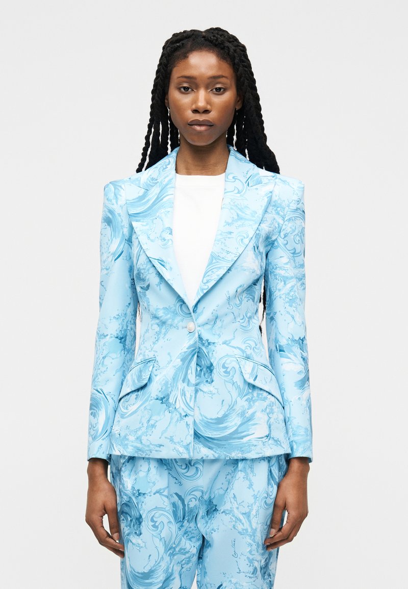 Γυναίκα με μακριά πλεξούδα που φοράει κομψό ανοιχτό μπλε κοστούμι με κυματιστά σχέδια και λευκό πουκάμισο, στέκεται μπροστά σε απλό φόντο.