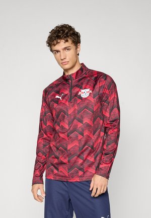 Rød og sort zigzag-mønstrede sportsjakke med høj krave og lynlås, der har et logo på brystet. Lavet af letvægtsmateriale.