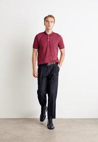 Burgundy poloko s krátkym rukávom, čierne skladané nohavice a čierne topánky. Model stojí na neutrálnom pozadí.