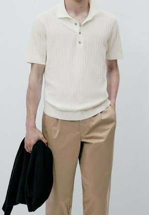 Man wearing a short-sleeve cream knit polo shirt and beige trousers, holding a black jacket in left hand, standing against a plain background.